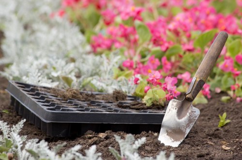 A close-up view of a garden bed during planting season, featuring rich, dark soil with a small metallic trowel that has a wooden handle embedded into the earth. Surrounding the planting area, there are vibrant pink and white flowering plants, possibly geraniums, with lush green leaves providing a colorful backdrop. A black seed tray, partially filled with soil, rests on the ground, indicating ongoing planting activity. The garden area appears well-maintained and organized, with the soil prepared for new plants. The lighting suggests natural daylight, and the scene is outdoors, likely in a well-kept front or back garden space in Clapham, typical of local residential landscaping and garden care activities conducted by Gardening Services Clapham.
